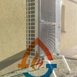 Zewnętrzna jednostka klimatyzacji Samsung zamontowana na białych wspornikach przy ścianie budynku o beżowej elewacji, widoczne logo firmy na dole zdjęcia.