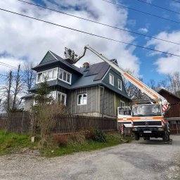 Topinstall - Montaż paneli słonecznych na dachu dwupiętrowego domu z wykorzystaniem podnośnika koszowego Skylift S 25, widoczne częściowo ocieplone ściany i drewniane ogrodzenie.