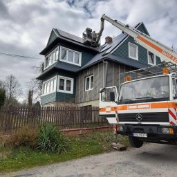 Topinstall - Montaż paneli słonecznych na dwupiętrowym domu z pomocą podnośnika Skylift S 24, widoczna elewacja ocieplona wełną mineralną, ogrodzenie drewniane, pochmurne niebo.