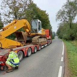 Topinstall - Żółta koparka na czerwonej platformie transportowej, zabezpieczana przez pracownika w odblaskowej kamizelce, na tle wiejskiej drogi i krajobrazu.