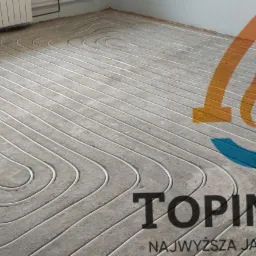 Białe rurki ogrzewania podłogowego ułożone w pętle na szarym podłożu, widoczny fragment grzejnika i logo firmy TOPINSTALL.
