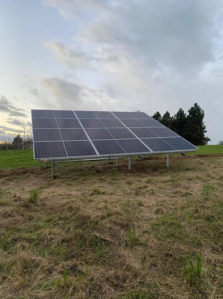 Panel słoneczny ustawiony na metalowej konstrukcji na nierównym terenie trawiastym, z drzewami w tle pod pochmurnym niebem.