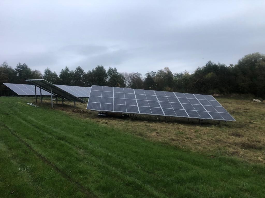 Rząd paneli fotowoltaicznych na metalowych stelażach, ustawionych na trawiastym wzgórzu z widokiem na las w pochmurny dzień.