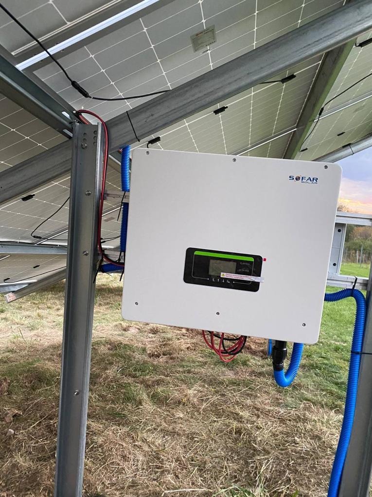 Biały inwerter solarny marki Sofar zamontowany na metalowej konstrukcji z panelami słonecznymi w tle, kable w niebieskich peszlach, widok z dołu na trawiastym terenie.