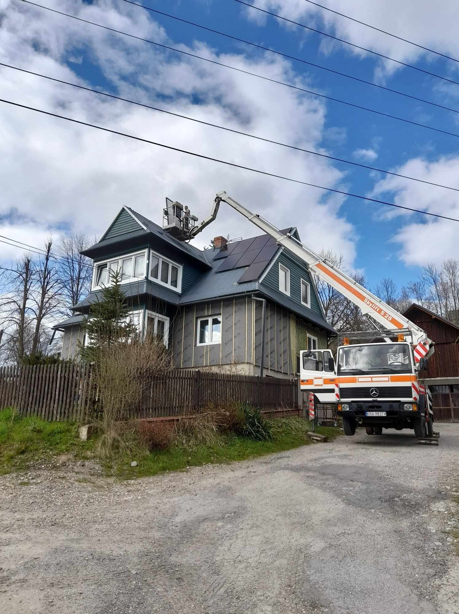 Montaż paneli słonecznych na dachu dwupiętrowego domu z wykorzystaniem podnośnika koszowego Skylift S 25, widoczne częściowo ocieplone ściany i drewniane ogrodzenie.
