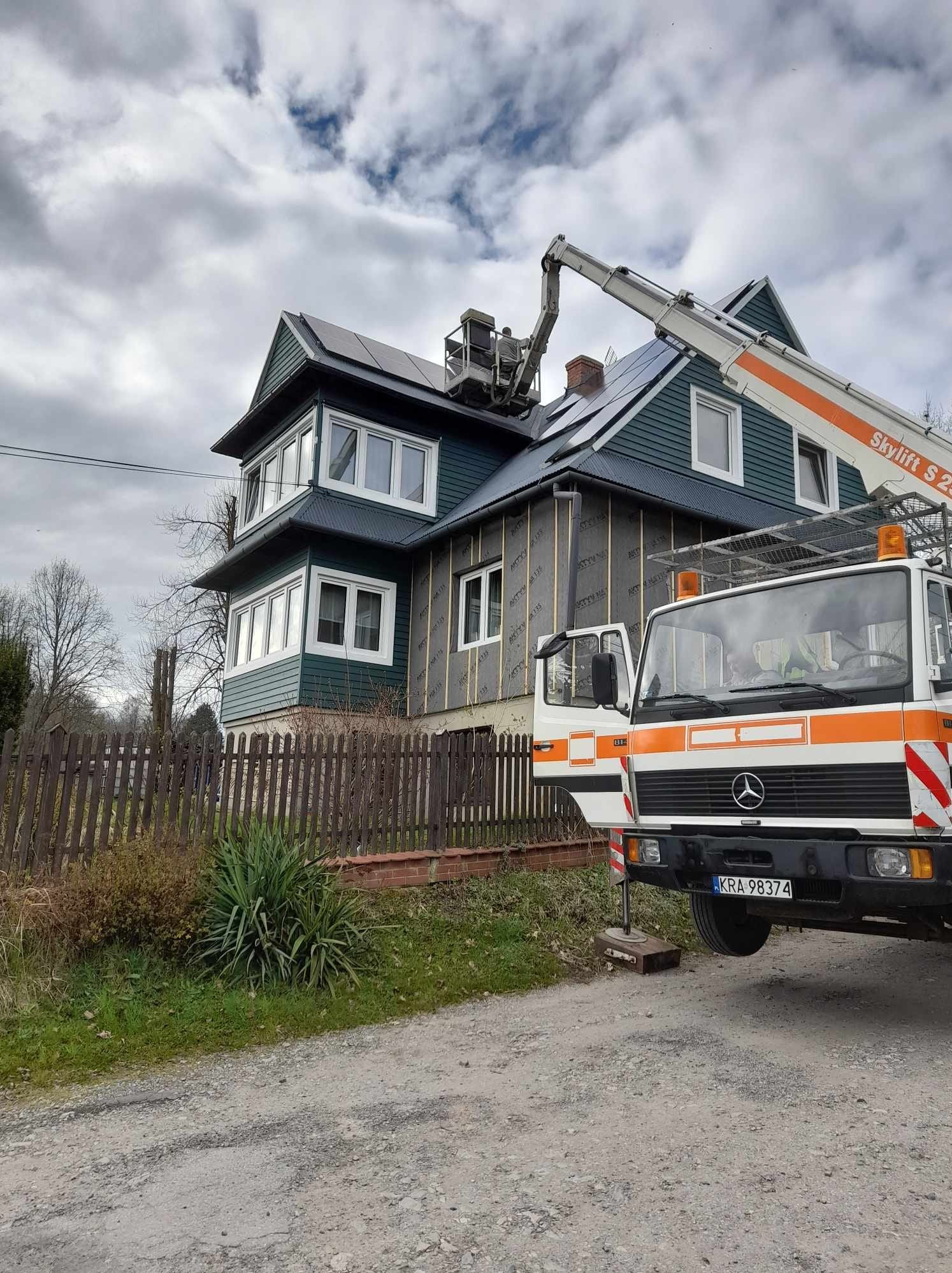 Montaż paneli słonecznych na dwupiętrowym domu z pomocą podnośnika Skylift S 24, widoczna elewacja ocieplona wełną mineralną, ogrodzenie drewniane, pochmurne niebo.