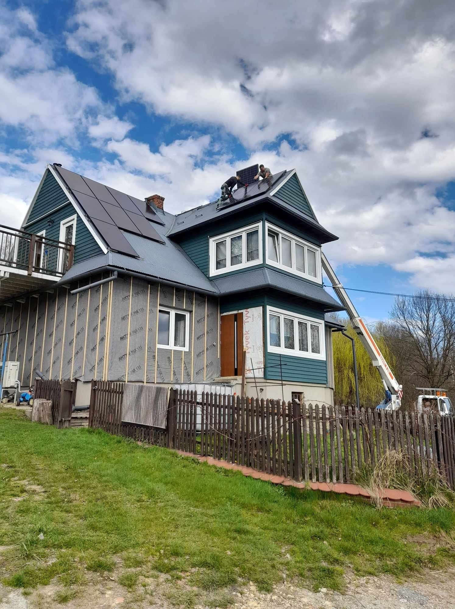 Montaż paneli fotowoltaicznych na dachu dwupiętrowego domu z podnośnika koszowego; widoczne ocieplenie ścian zewnętrznych budynku.