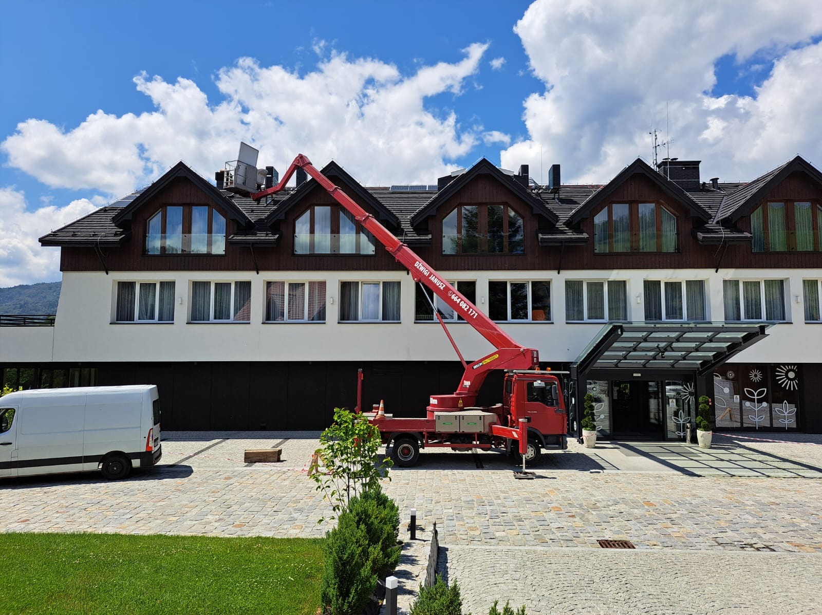 Czerwony dźwig samochodowy z podnośnikiem teleskopowym unosi element instalacji na dach trzypiętrowego budynku w słoneczny dzień z dekoracyjnymi oknami i wejściem.