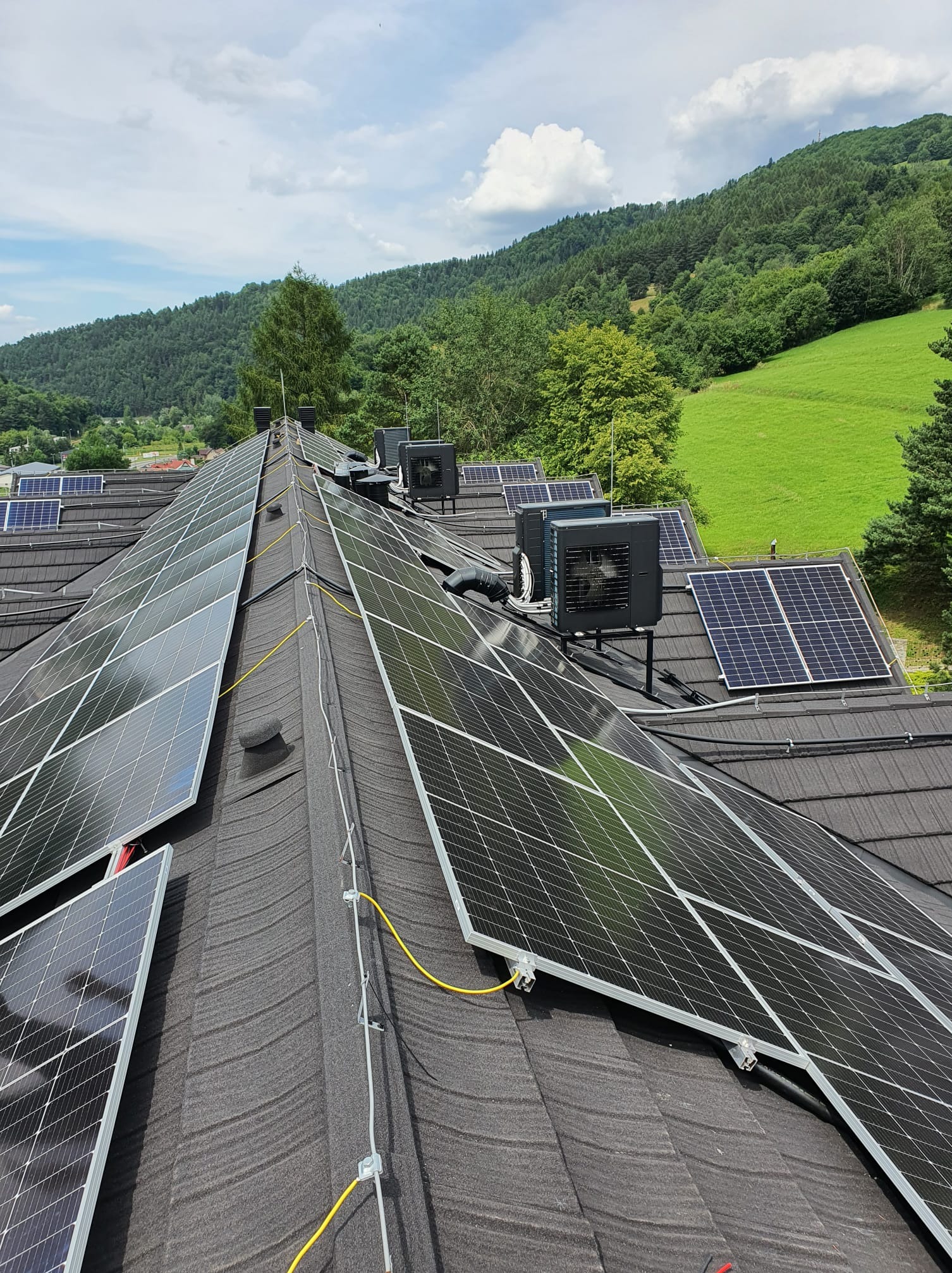 Panele słoneczne i jednostki zewnętrzne na dachu domu z widokiem na zielone wzgórza w oddali.