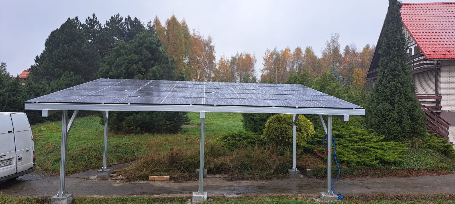 Zadaszenie z paneli fotowoltaicznych na metalowej konstrukcji w Myślenicach, widok z boku na tle zieleni i domu z czerwoną dachówką.