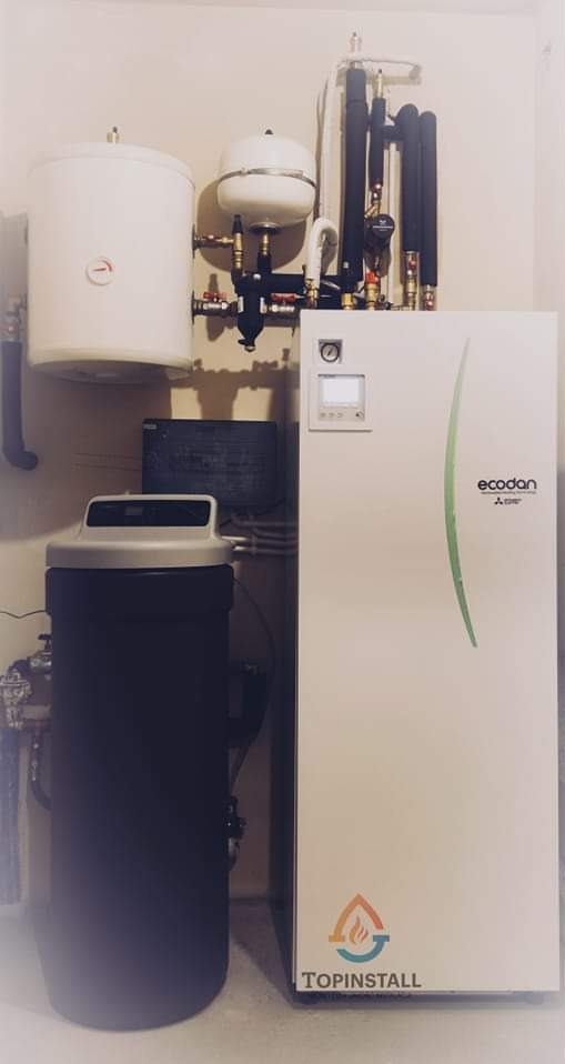 Biała pompa ciepła Ecodan Mitsubishi z zbiornikiem buforowym i zmiękczaczem wody w kotłowni, widoczne rury instalacyjne oraz logo firmy Topinstall.