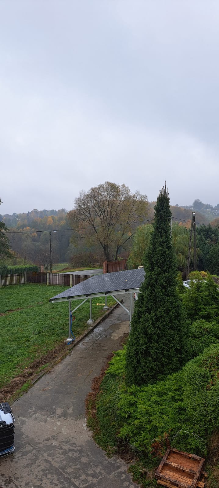 Zadaszenie parkingu z panelami fotowoltaicznymi w jesiennej scenerii, widok z góry na posesję z zielonym trawnikiem i drzewami w tle, pochmurny dzień.