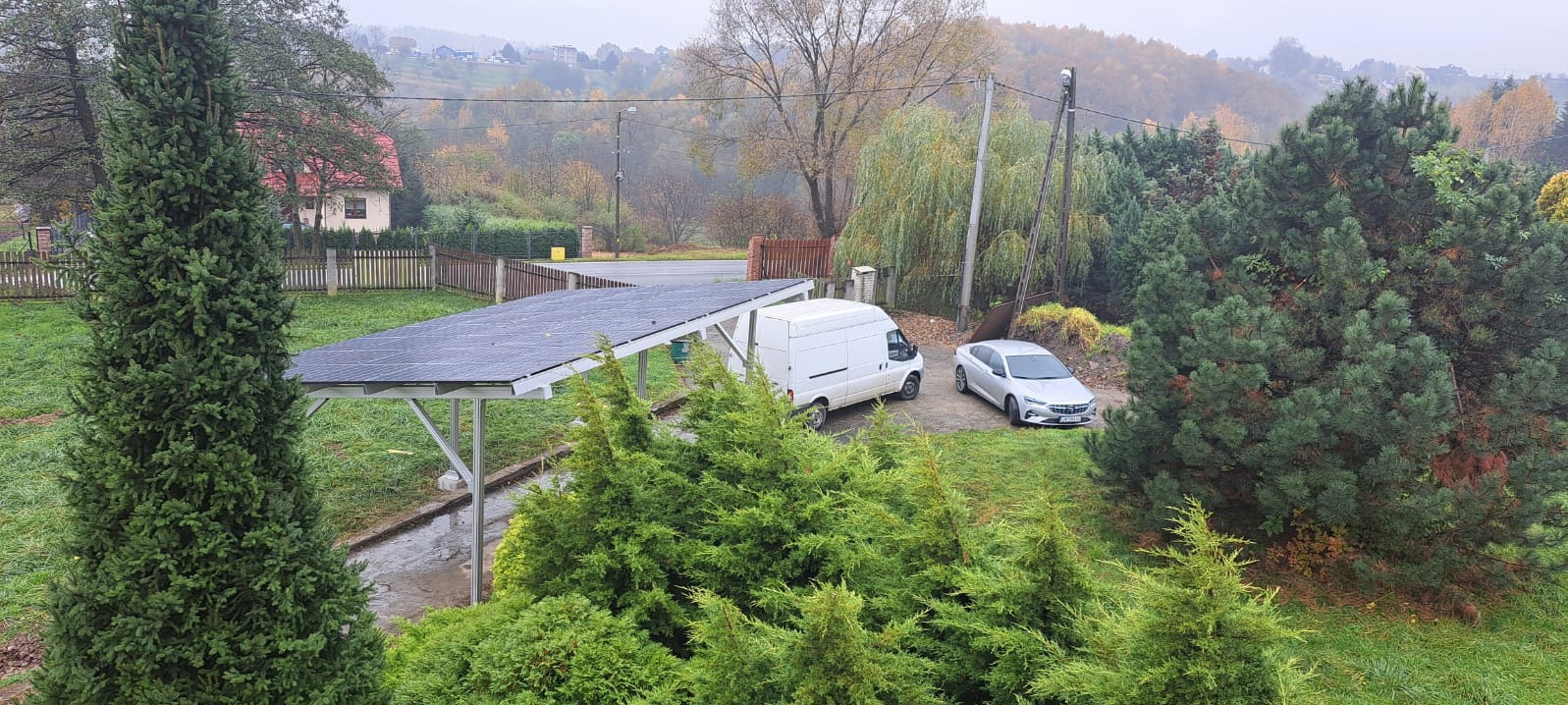 Zewnętrzna instalacja fotowoltaiczna na konstrukcji w formie wiaty, obok zaparkowany biały bus i srebrny samochód osobowy, widok z góry w pochmurny dzień.