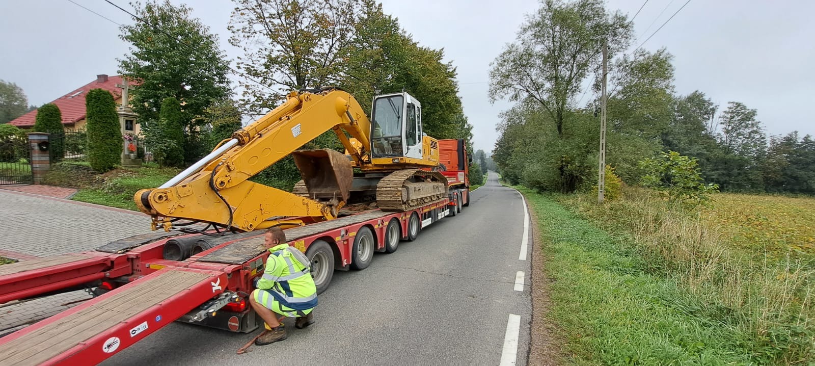 Żółta koparka na czerwonej platformie transportowej, zabezpieczana przez pracownika w odblaskowej kamizelce, na tle wiejskiej drogi i krajobrazu.