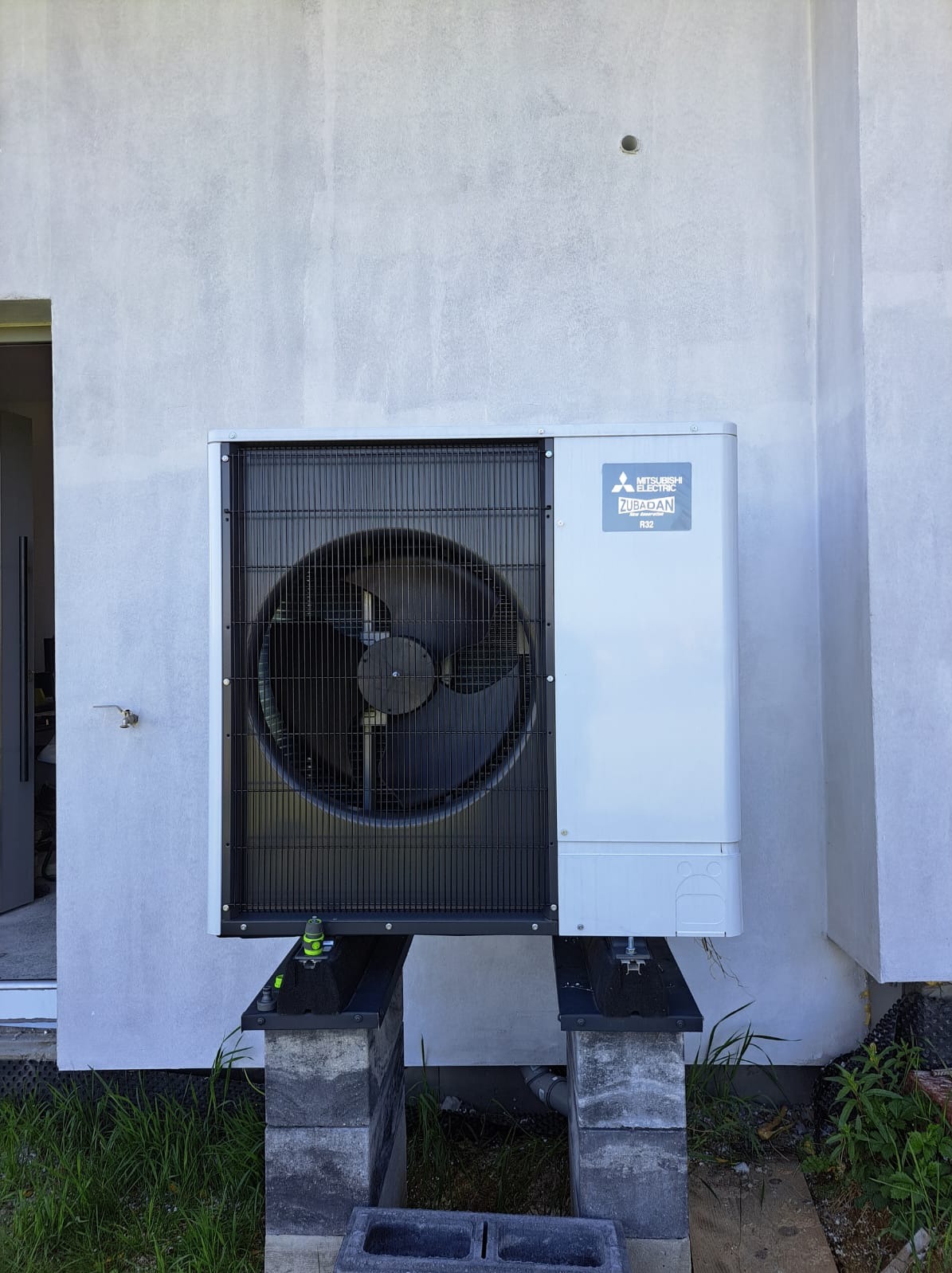 Zewnętrzna jednostka pompy ciepła Mitsubishi Zubadan zamontowana na betonowych blokach przy szarej ścianie budynku z widocznym wejściem.