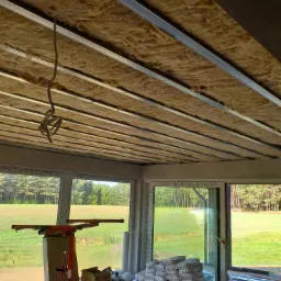 Montaż izolacji z wełny mineralnej na suficie podwieszanym, widoczne profile metalowe, worki z zaprawą i podnośnik do płyt gipsowych w pomieszczeniu z dużymi oknami z widokiem na zieleń.