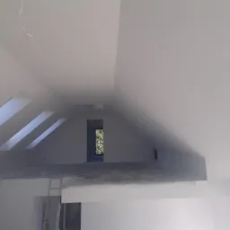 Poddasze w trakcie wykańczania: widoczne skosy, okna dachowe, ściany częściowo zagruntowane, częściowo pomalowane na biało, drabina oparta o ścianę, widok na zieleń przez okno.