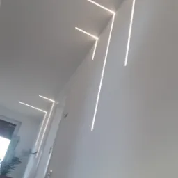 Nowoczesne oświetlenie sufitowe z liniowymi lampami LED w kształcie zygzaków, widok na schody i roślinę w tle.