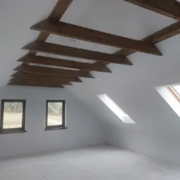 Poddasze z drewnianymi belkami stropowymi, dwoma oknami fasadowymi i dwoma połaciowymi, świeżo po wykonaniu gładzi i przygotowaniu do dalszych prac wykończeniowych.