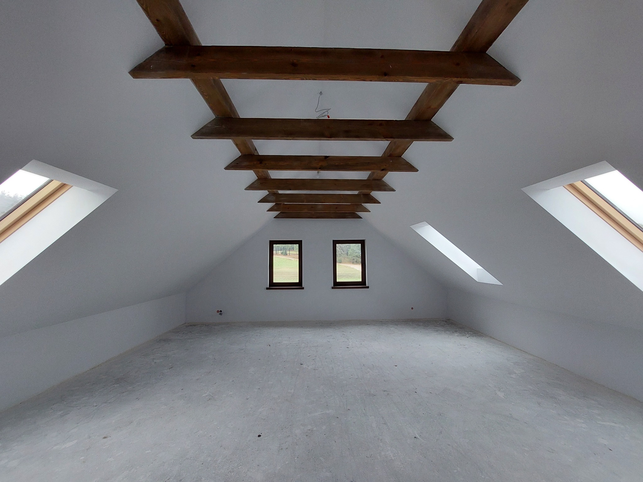 Poddasze po szpachlowaniu, z widocznymi drewnianymi belkami stropowymi, oknami dachowymi i dwoma oknami szczytowymi. Powierzchnia podłogi szara, gotowa do dalszych prac wykończeniowych.