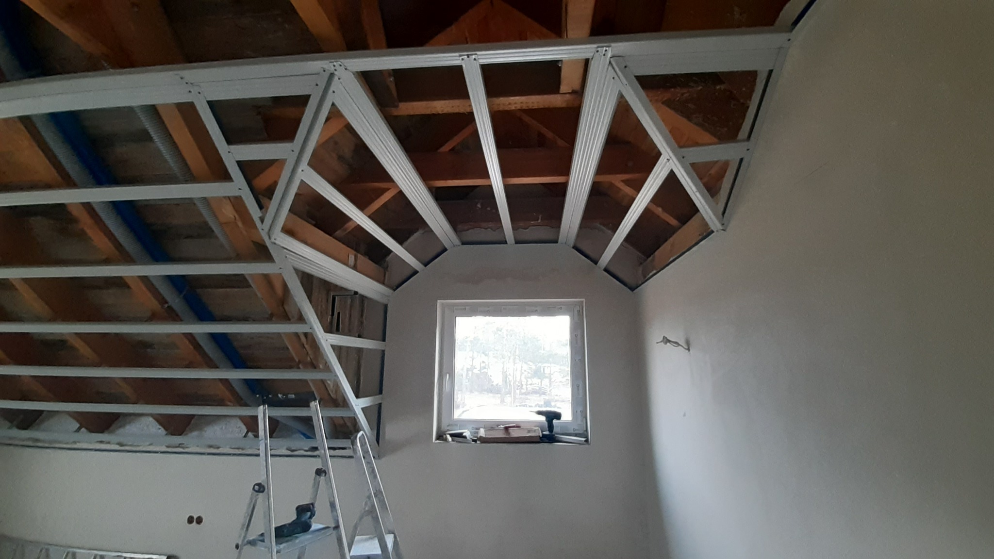 Konstrukcja metalowa sufitu podwieszanego na poddaszu, widoczne drewniane elementy więźby dachowej, okno z narzędziami na parapecie, drabina przystawna w rogu pomieszczenia.