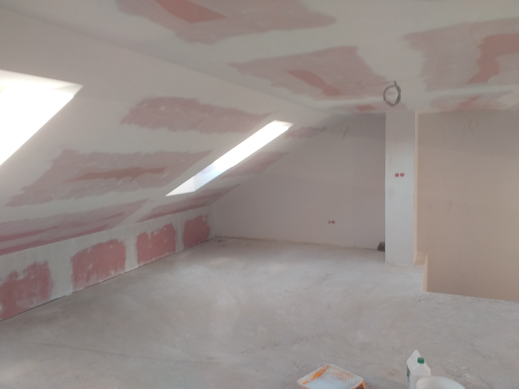 Poddasze w trakcie przygotowania do malowania, widoczne fragmenty ścian i sufitu z naniesioną warstwą szpachli, okna dachowe wpuszczające światło, na podłodze wiaderka z farbą i kuweta malarska.