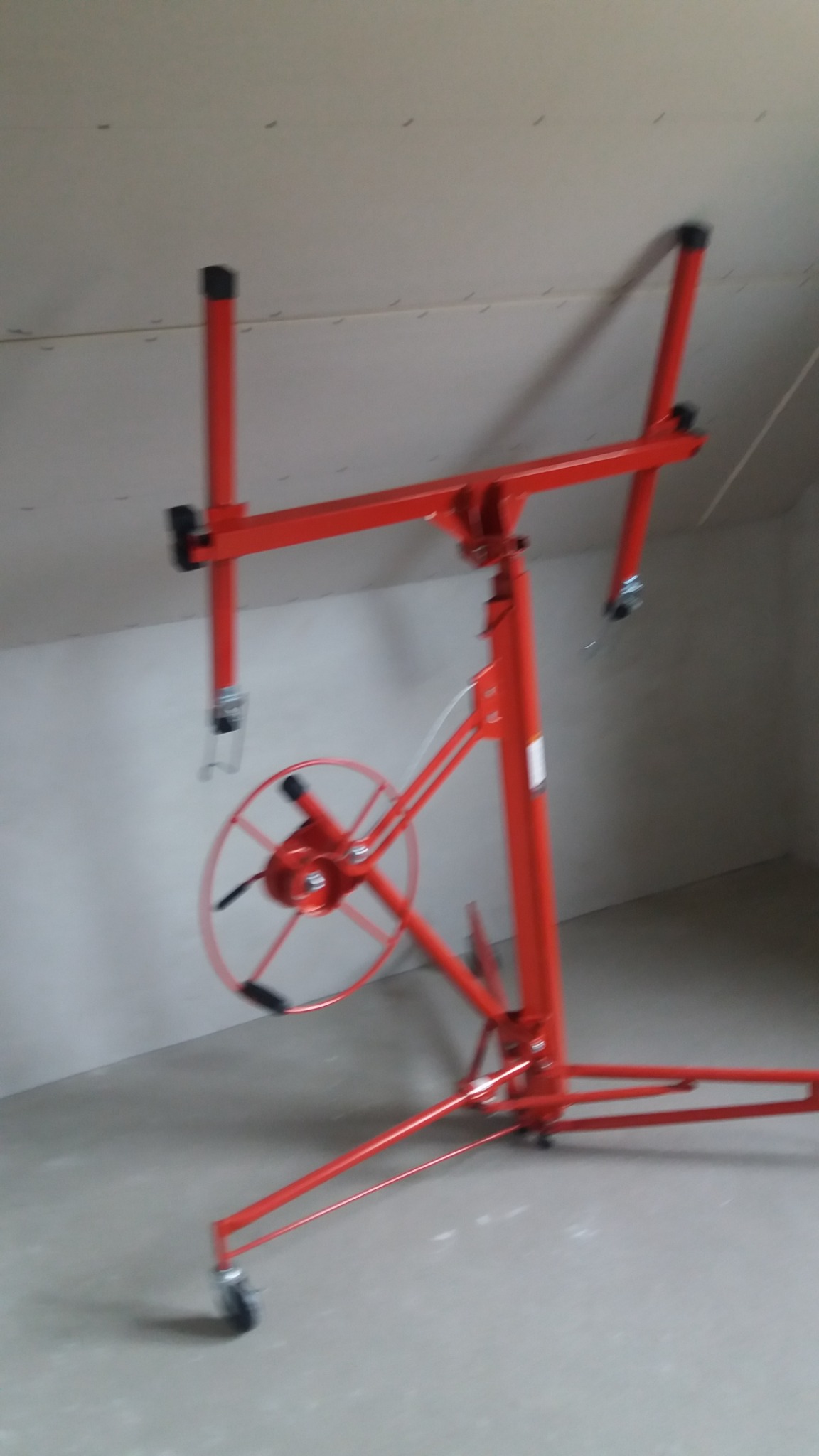 Czerwony podnośnik do płyt gipsowo-kartonowych ustawiony w pomieszczeniu ze skośnym sufitem, widoczne łączenia płyt na suficie i ścianach.