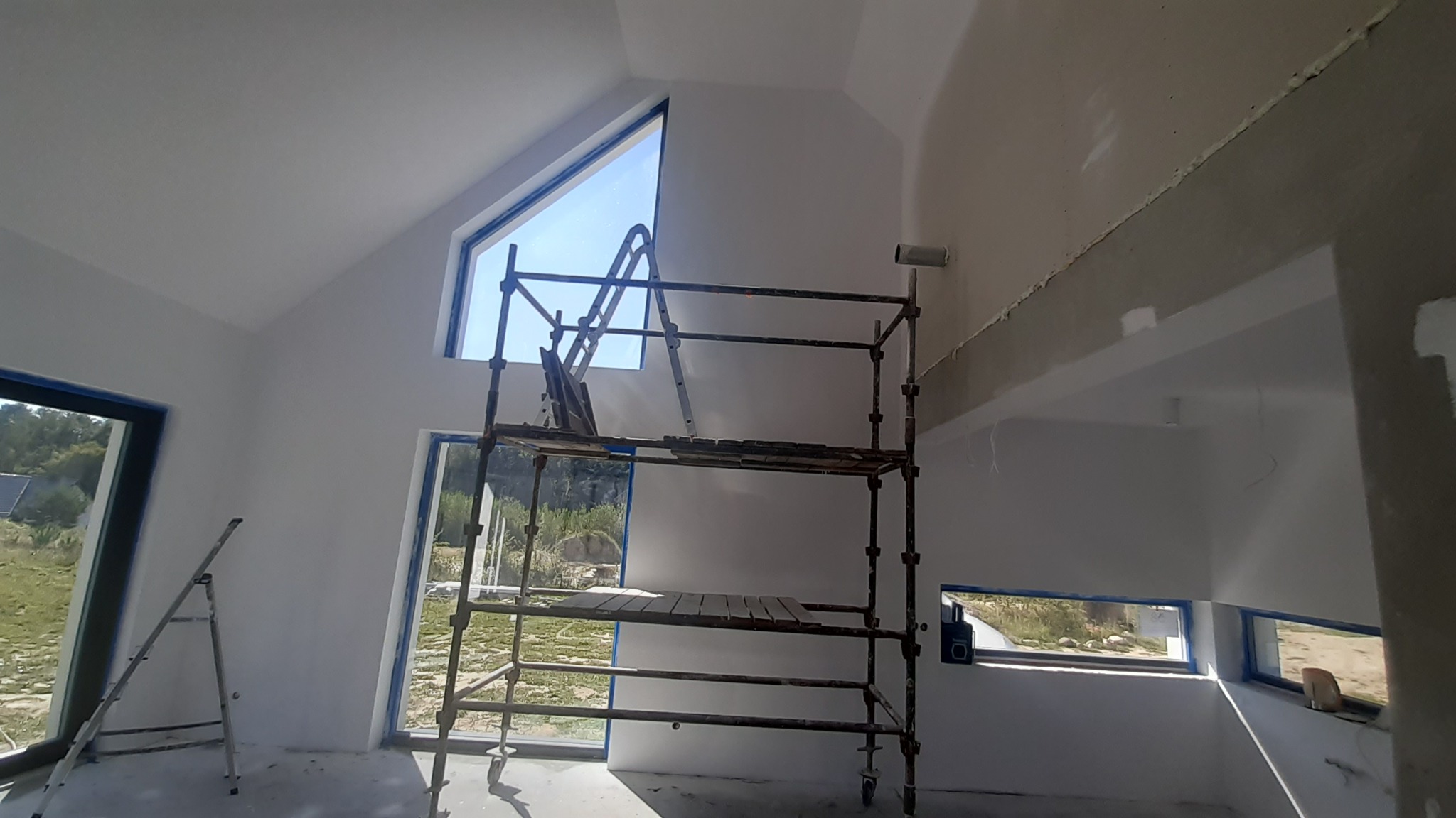 Wnętrze w trakcie prac wykończeniowych: szpachlowane ściany, zabezpieczone okna taśmą malarską, rusztowanie i drabina w pomieszczeniu z oknami o nietypowym kształcie.