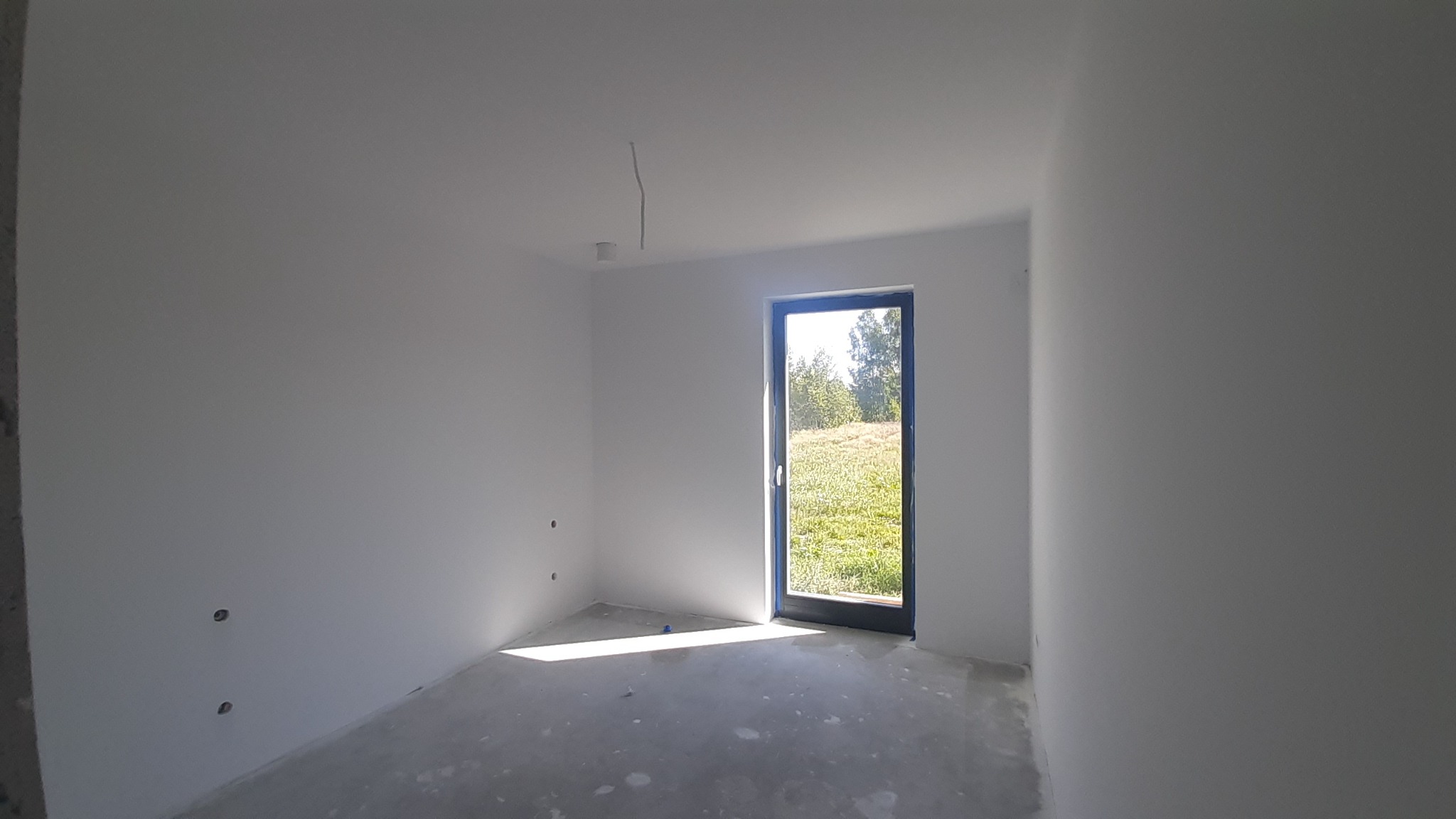 Wnętrze świeżo wykończonego pokoju z gładkimi, białymi ścianami, betonową podłogą i oknem z widokiem na zieleń, z widocznymi śladami po gniazdkach elektrycznych.
