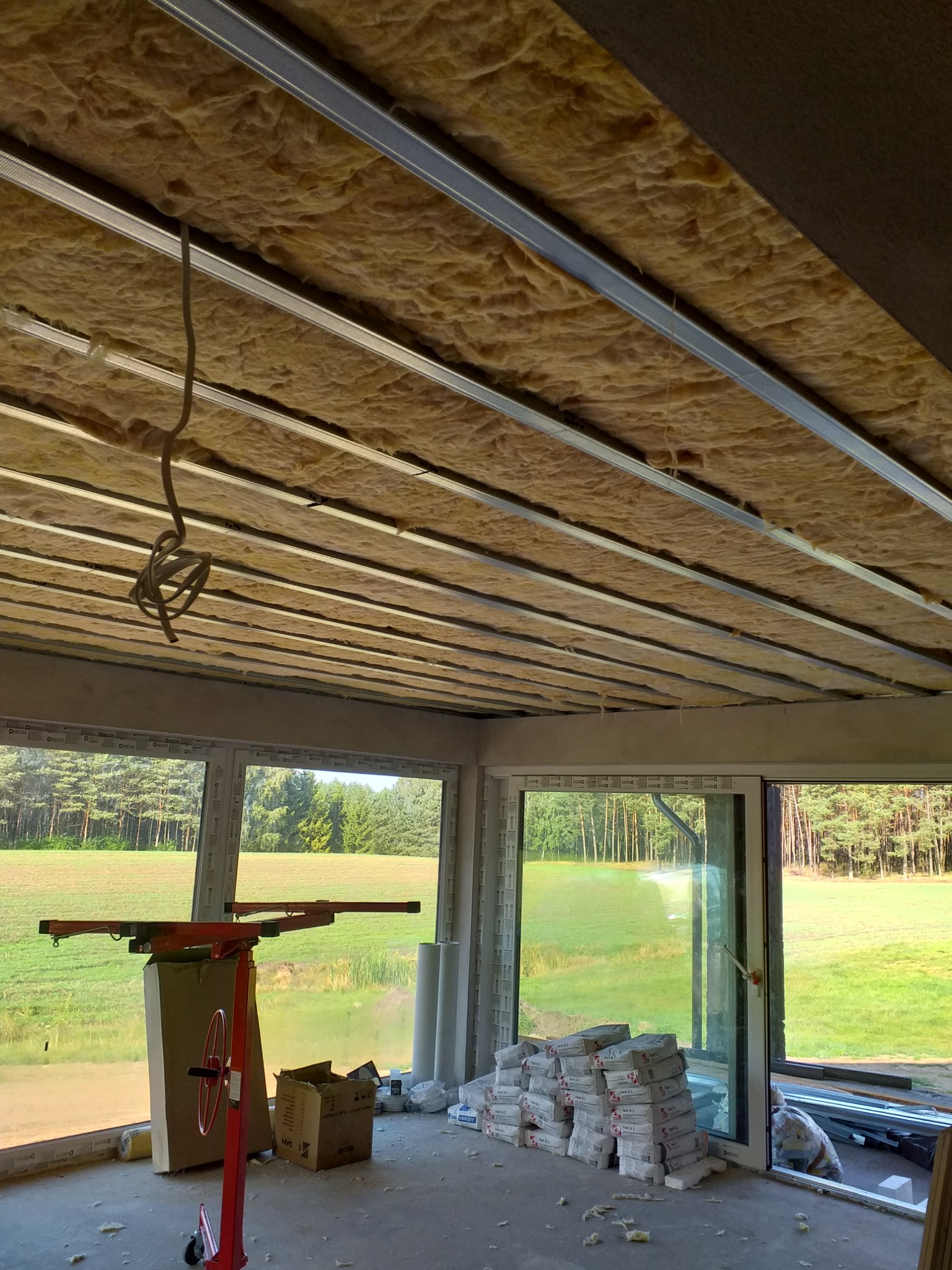 Montaż izolacji z wełny mineralnej na suficie podwieszanym, widoczne profile metalowe, worki z zaprawą i podnośnik do płyt gipsowych w pomieszczeniu z dużymi oknami z widokiem na zieleń.