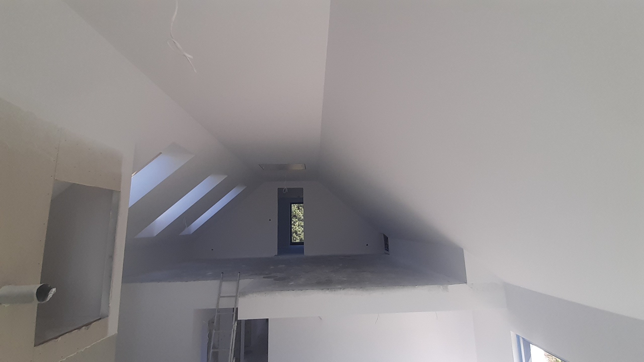 Poddasze w trakcie wykańczania: widoczne skosy, okna dachowe, ściany częściowo zagruntowane, częściowo pomalowane na biało, drabina oparta o ścianę, widok na zieleń przez okno.
