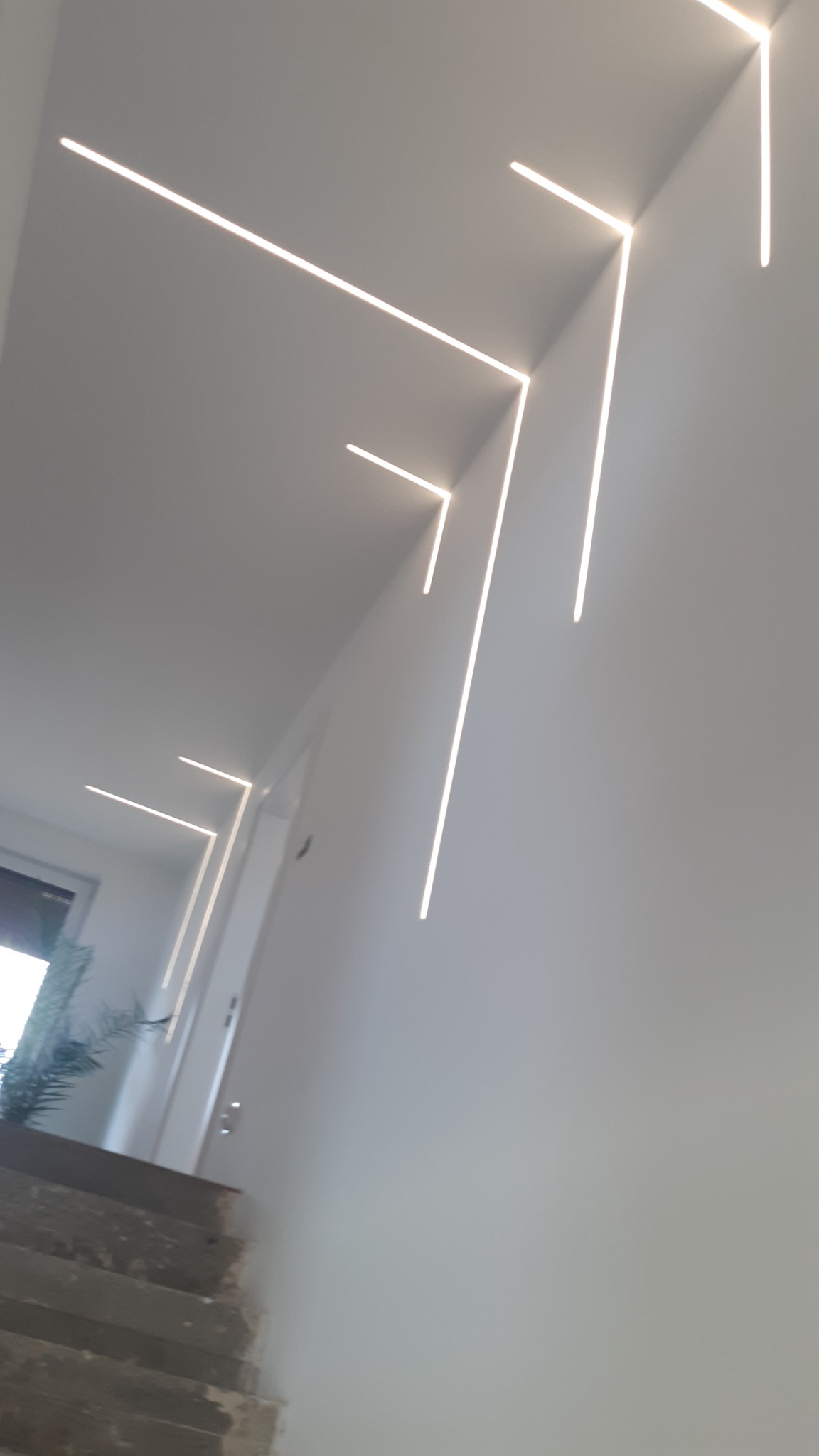 Nowoczesne oświetlenie sufitowe z liniowymi lampami LED w kształcie zygzaków, widok na schody i roślinę w tle.