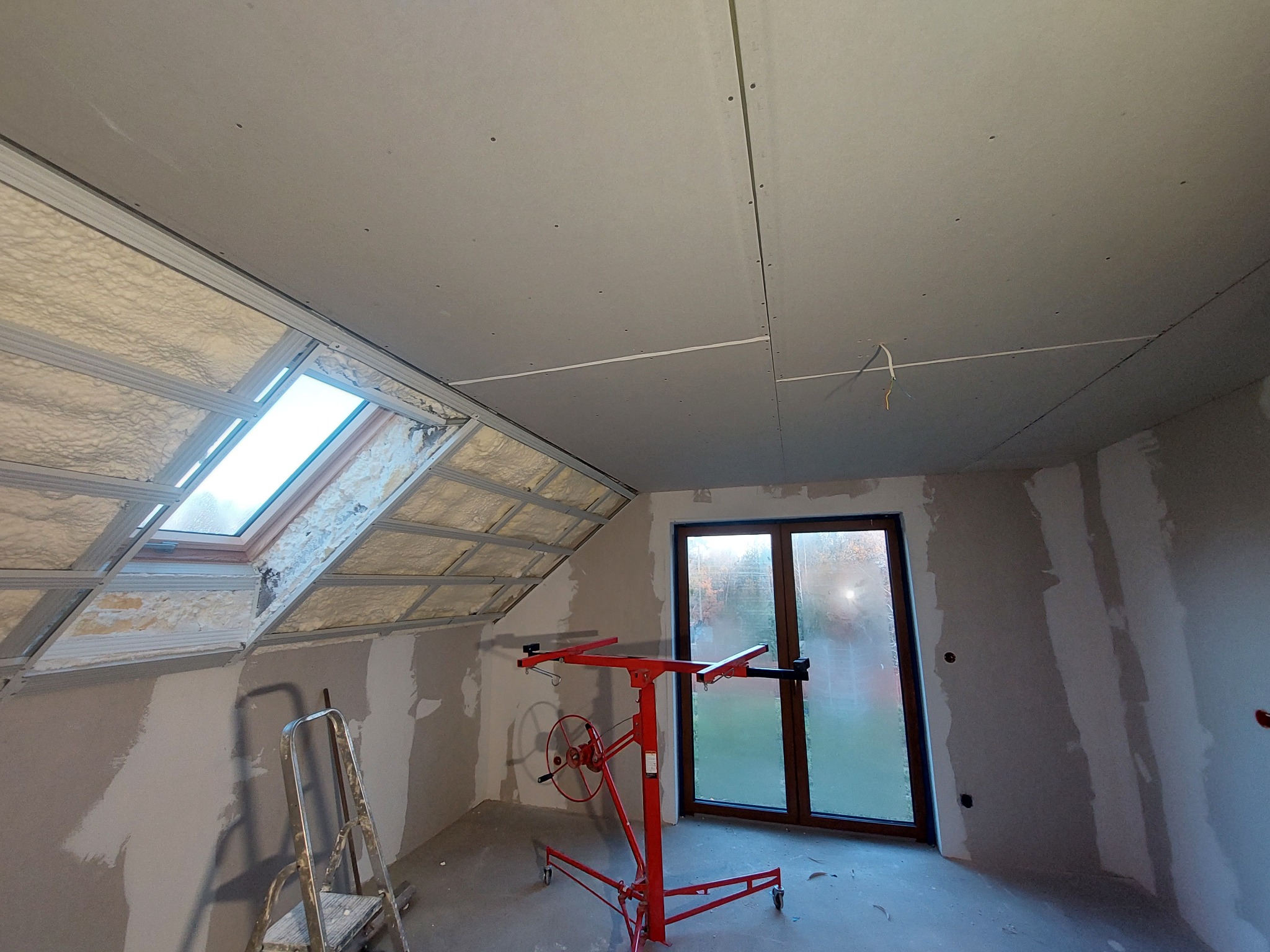 Pokój na poddaszu w trakcie wykańczania: zamontowane płyty gipsowo-kartonowe na suficie i skosach, widoczna wełna mineralna pod płytami, czerwony podnośnik do płyt g-k, drabina aluminiowa i okno...