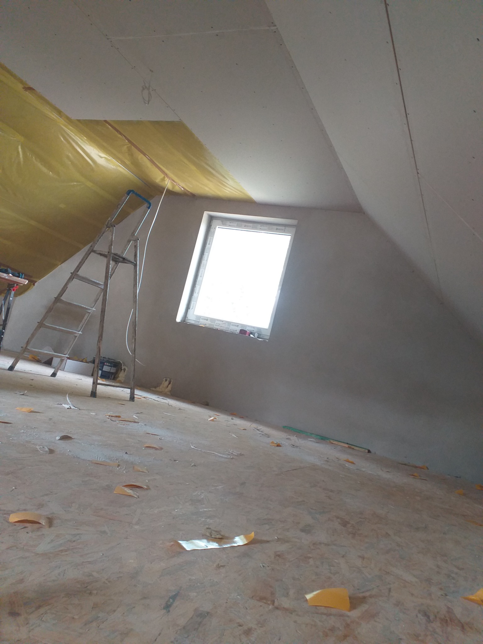 Poddasze w trakcie prac wykończeniowych: ocieplona skośna ściana, okno, drabina i surowy parkiet z resztkami taśmy malarskiej na podłodze.