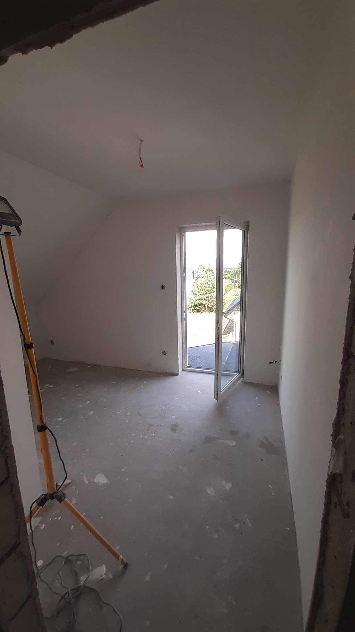 Pokój na poddaszu w trakcie wykańczania, ściany przygotowane pod malowanie, podłoga z śladami po szpachlowaniu, otwarte drzwi balkonowe wpuszczające światło dzienne, lampa budowlana na trójnogu...