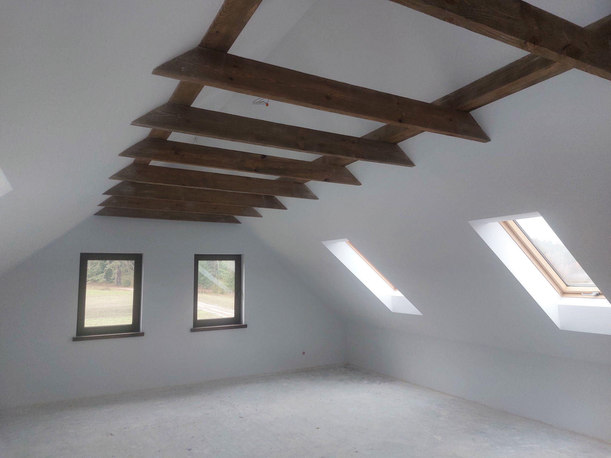 Poddasze z drewnianymi belkami stropowymi, dwoma oknami fasadowymi i dwoma połaciowymi, świeżo po wykonaniu gładzi i przygotowaniu do dalszych prac wykończeniowych.