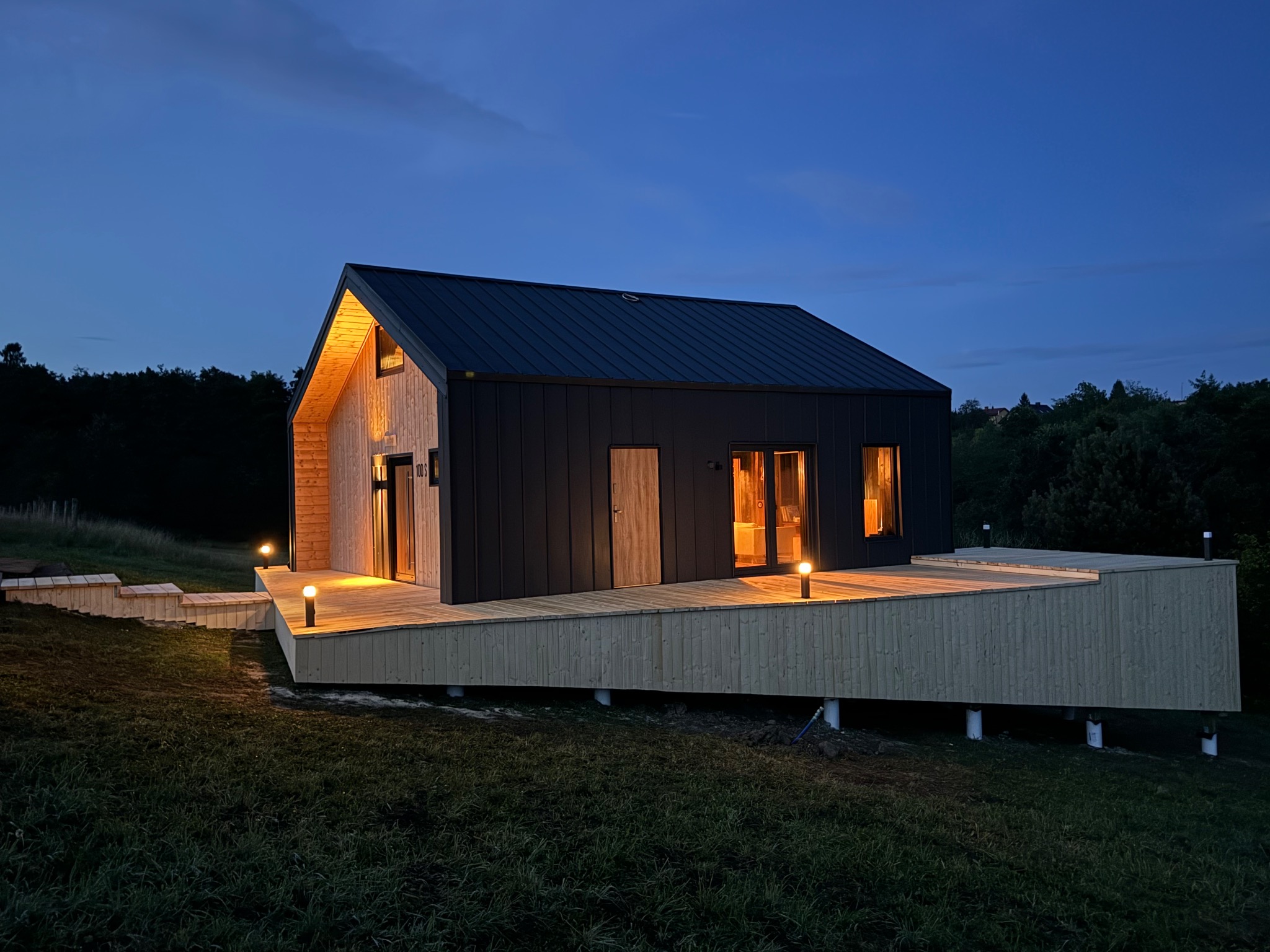 Nowoczesny dom szkieletowy z ciemną blachą na dachu i jasną drewnianą elewacją, podświetlony wieczornym światłem, z tarasem na podwyższeniu.