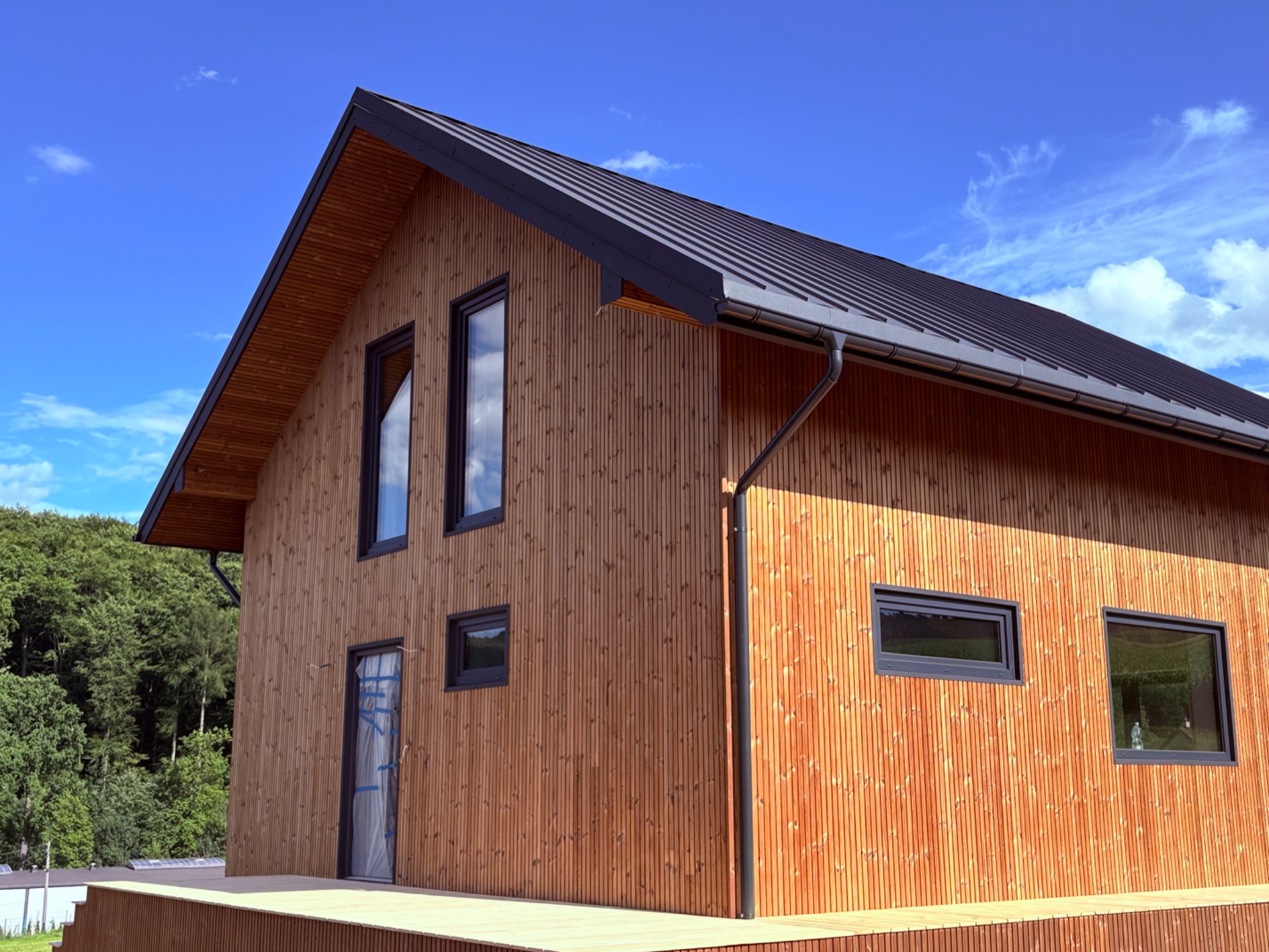 Nowoczesny dom z drewnianą elewacją w pionowe panele, czarny dach i okna. Widok z zewnątrz, błękitne niebo w tle. Drzwi wejściowe zabezpieczone folią.