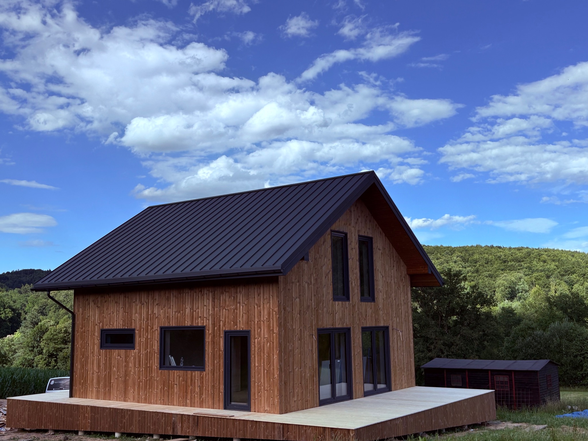 Nowoczesny dom szkieletowy z drewnianą elewacją i ciemnym dachem blachodachówką na tle błękitnego nieba z obłokami i zielonego lasu. Widoczny taras.