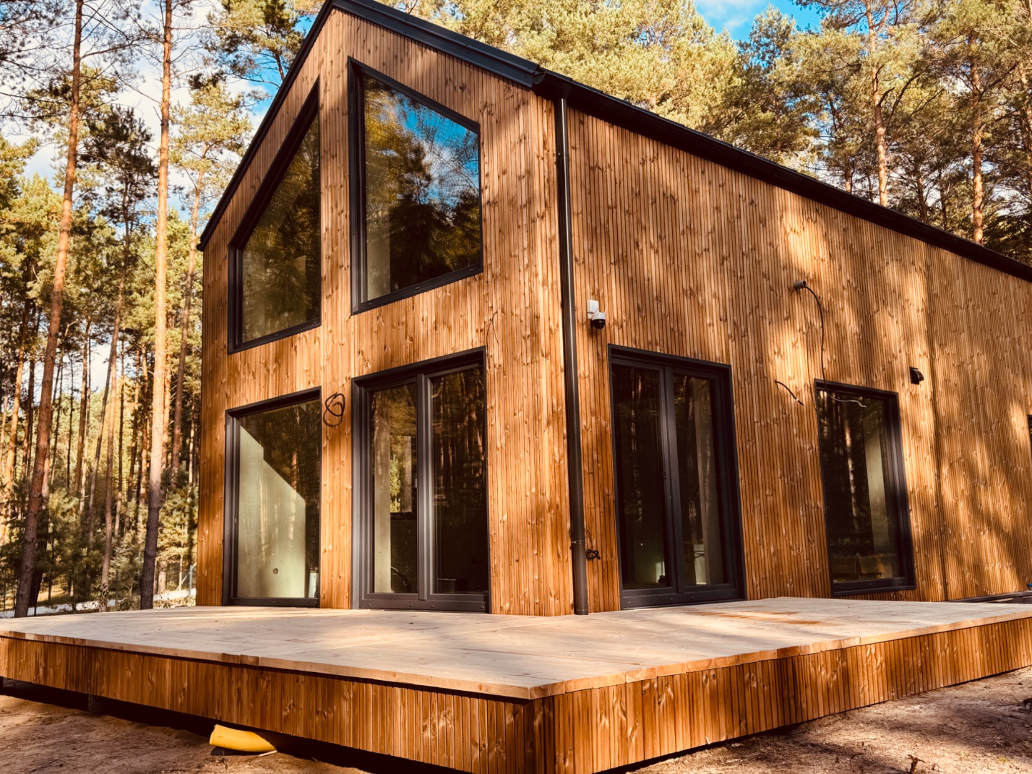 Nowoczesny dom szkieletowy z drewnianą elewacją i dużymi oknami, otoczony lasem. Widoczny taras z desek i zainstalowany system monitoringu.