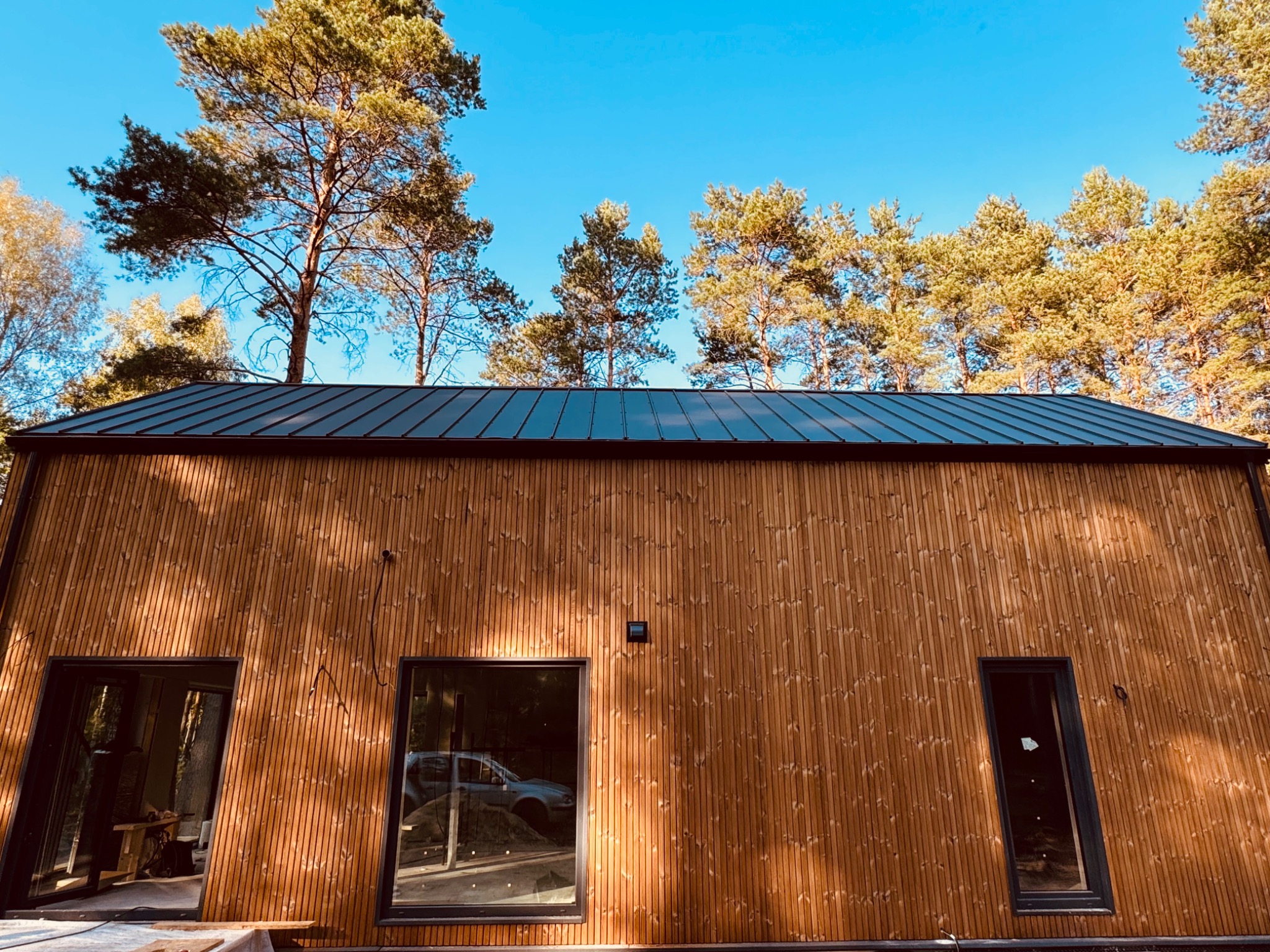 Nowoczesny dom z drewnianą elewacją i ciemnym dachem na tle lasu i błękitnego nieba. Widoczne okna z refleksami i częściowo odsłonięte wnętrze.