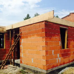 Budowa domu jednorodzinnego w stanie surowym otwartym, widoczne ściany z czerwonej cegły, drewniane elementy konstrukcyjne dachu i podpory, oraz prowizoryczna drewniana drabina oparta o ścianę.