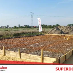 Zbrojenie fundamentu w drewnianym szalunku, widok z góry na plac budowy z hałdą ziemi i słupem wysokiego napięcia w tle.