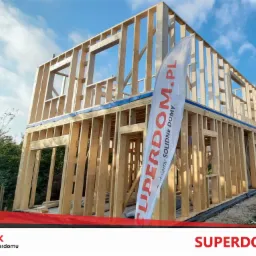 Dwukondygnacyjny budynek w trakcie budowy, konstrukcja szkieletowa z drewnianych belek, widoczny fundament i flaga reklamowa firmy.