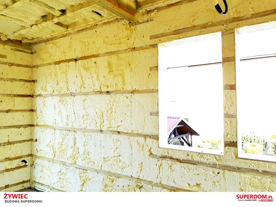Wnętrze domu w trakcie budowy, ściany i sufit pokryte żółtą pianką izolacyjną, widoczne drewniane elementy konstrukcyjne i dwa otwory okienne z widokiem na zewnątrz.