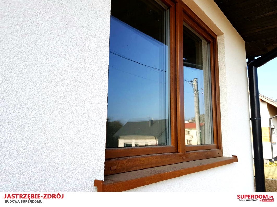 Fragment ściany budynku z oknem o drewnianej ramie, w szybie odbija się sąsiedni dom i niebo.