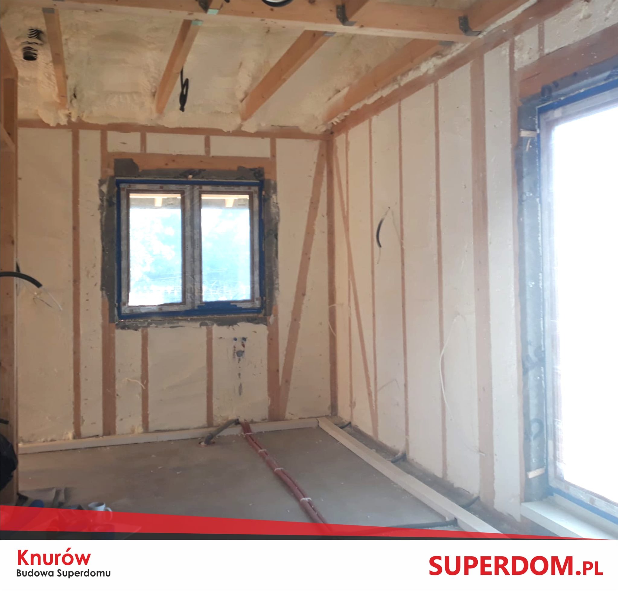 Wnętrze w trakcie budowy: ściany z drewnianą konstrukcją, wypełnione pianą izolacyjną, okno z nieotynkowanym obramowaniem, widoczne instalacje podłogowe.
