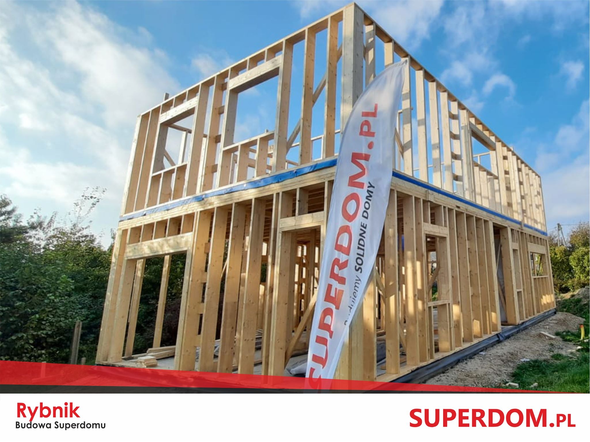 Dwukondygnacyjny budynek w trakcie budowy, konstrukcja szkieletowa z drewnianych belek, widoczny fundament i flaga reklamowa firmy.