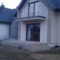 Kosmetal Adam Malkowski - Dom z balkonem i balustradą ze stali nierdzewnej. Widoczne betonowe schody wejściowe i okna. Tło stanowią sąsiednie budynki i niebo.