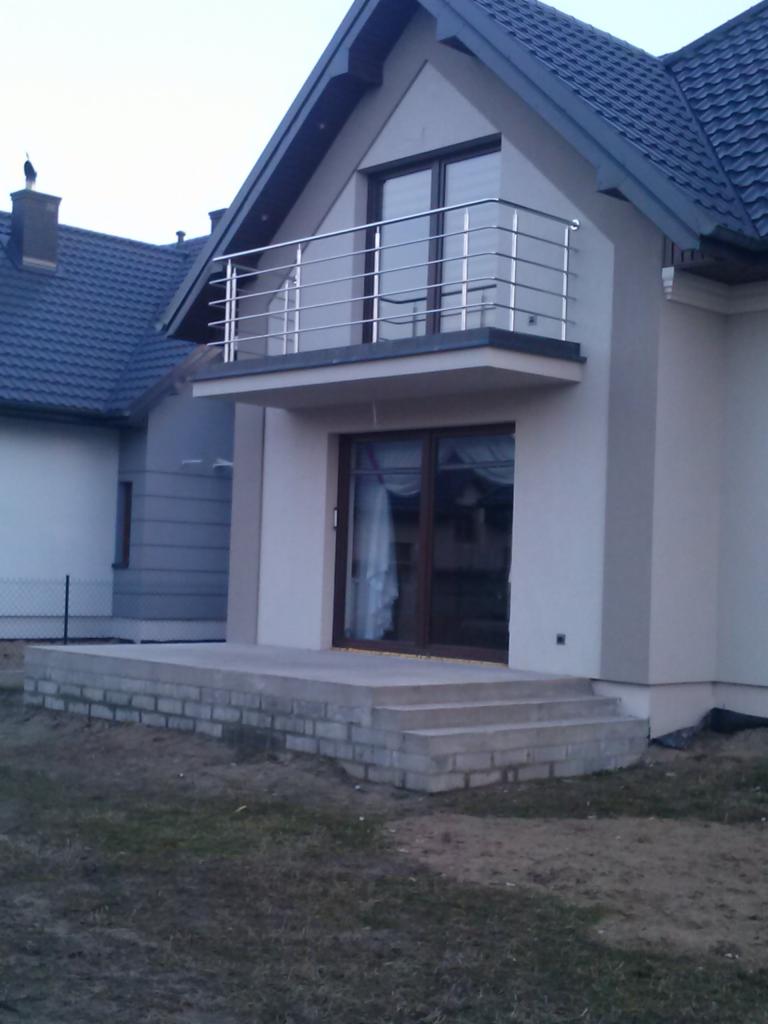 Dom z balkonem i balustradą ze stali nierdzewnej. Widoczne betonowe schody wejściowe i okna. Tło stanowią sąsiednie budynki i niebo.